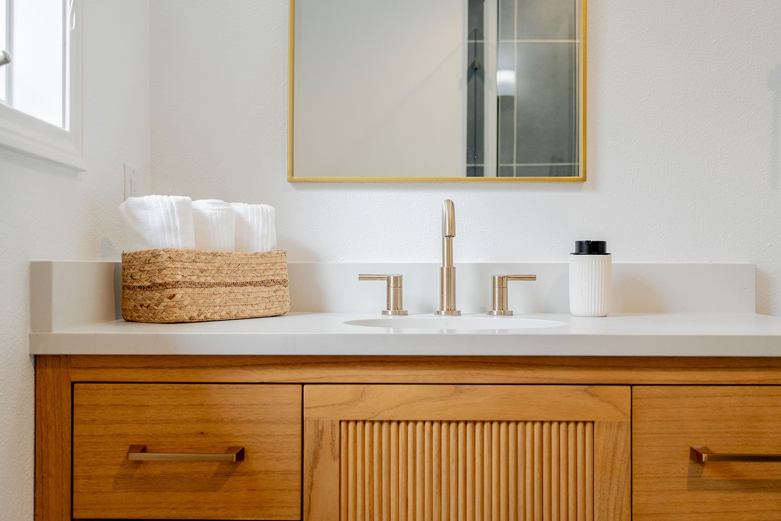 Modern bathroom vanity featuring a light wood cabinet, white countertop, brushed gold faucet, and rectangular gold mirror.