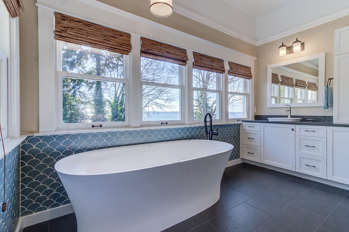 Primary bathroom remodel featuring a white freestanding soaking tub, blue scallop tile accent wall, and woven window shades.