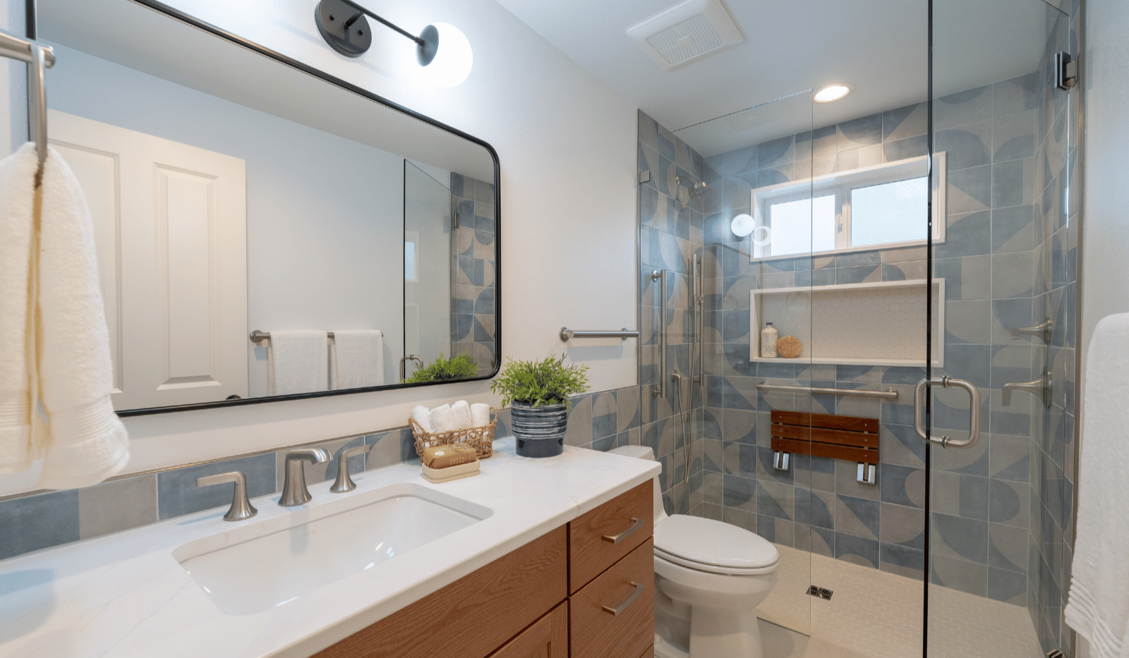 West Seattle accessible guest bathroom remodel with blue geometric shower tile, frameless glass, grab bars, fold-down shower seat, and wood vanity.